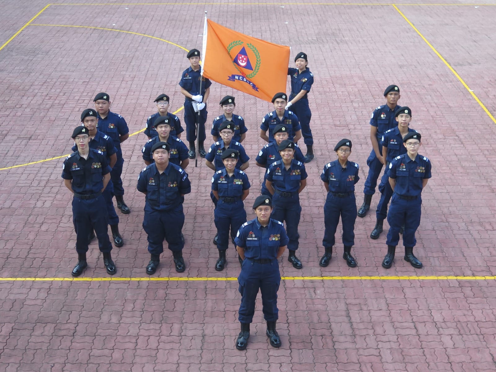 A group of youth cadets in navy uniforms and black berets stand in formation holding an orange NCDCC flag on a paved courtyard.